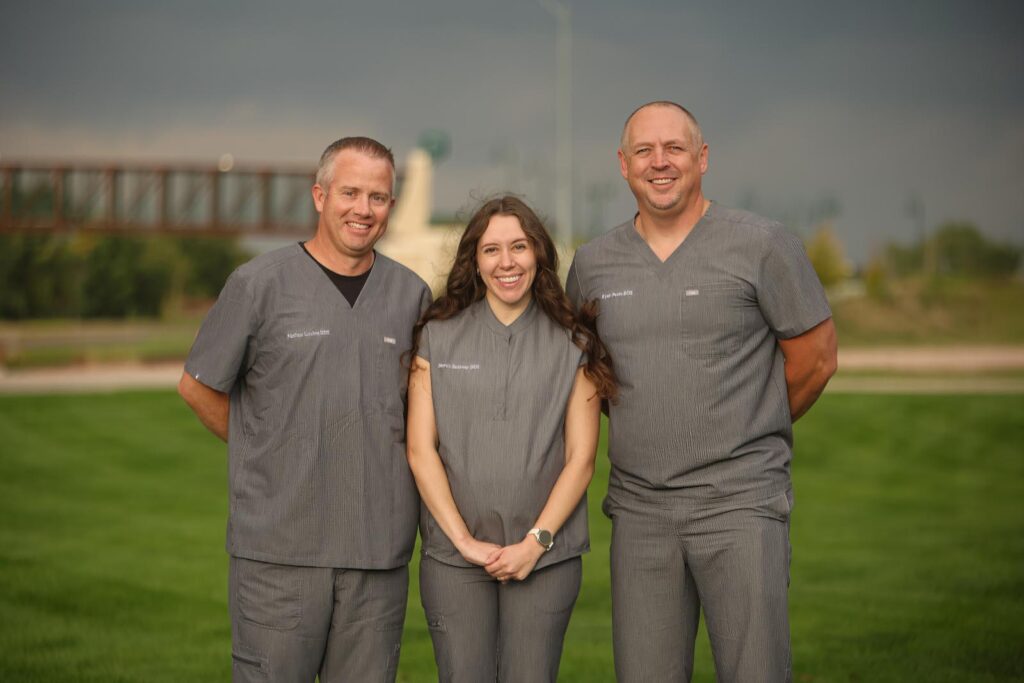 Three Platte Valley Dental Group doctors standing together outdoors in matching gray scrubs, smiling on a grassy lawn with a bridge and monument in the background.