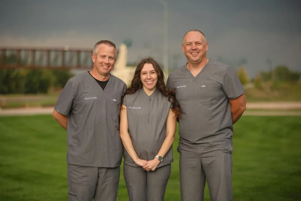 Three Platte Valley Dental Group doctors standing together outdoors in matching gray scrubs, smiling on a grassy lawn with a bridge and monument in the background.