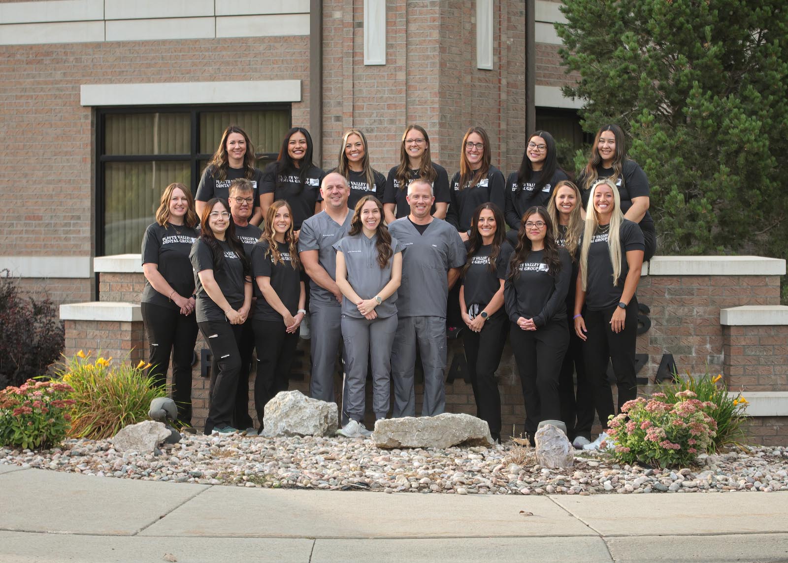 Group photo of Platte Valley Dental Group team standing outside the clinic, featuring 18 staff members in matching black 'Sage Dental' shirts and gray scrubs, posed in front of a brick building with landscaped flowers and shrubs.